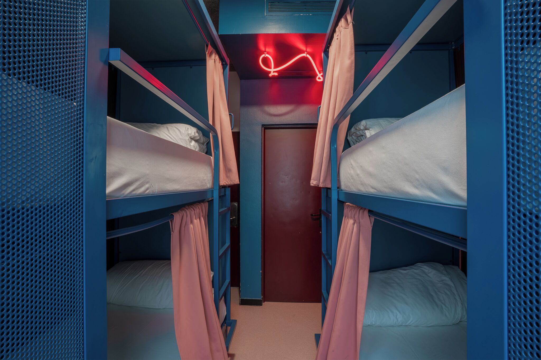 Een slaapkamer met stapelbedden in het blauw, roze gordijnen en een neonbord aan het plafond.