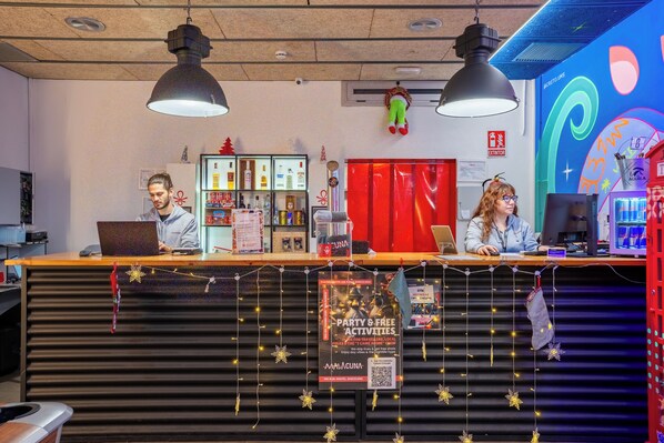 Un mostrador de recepción con dos empleados, un refrigerador lleno de bebidas y un mural de pared colorido.