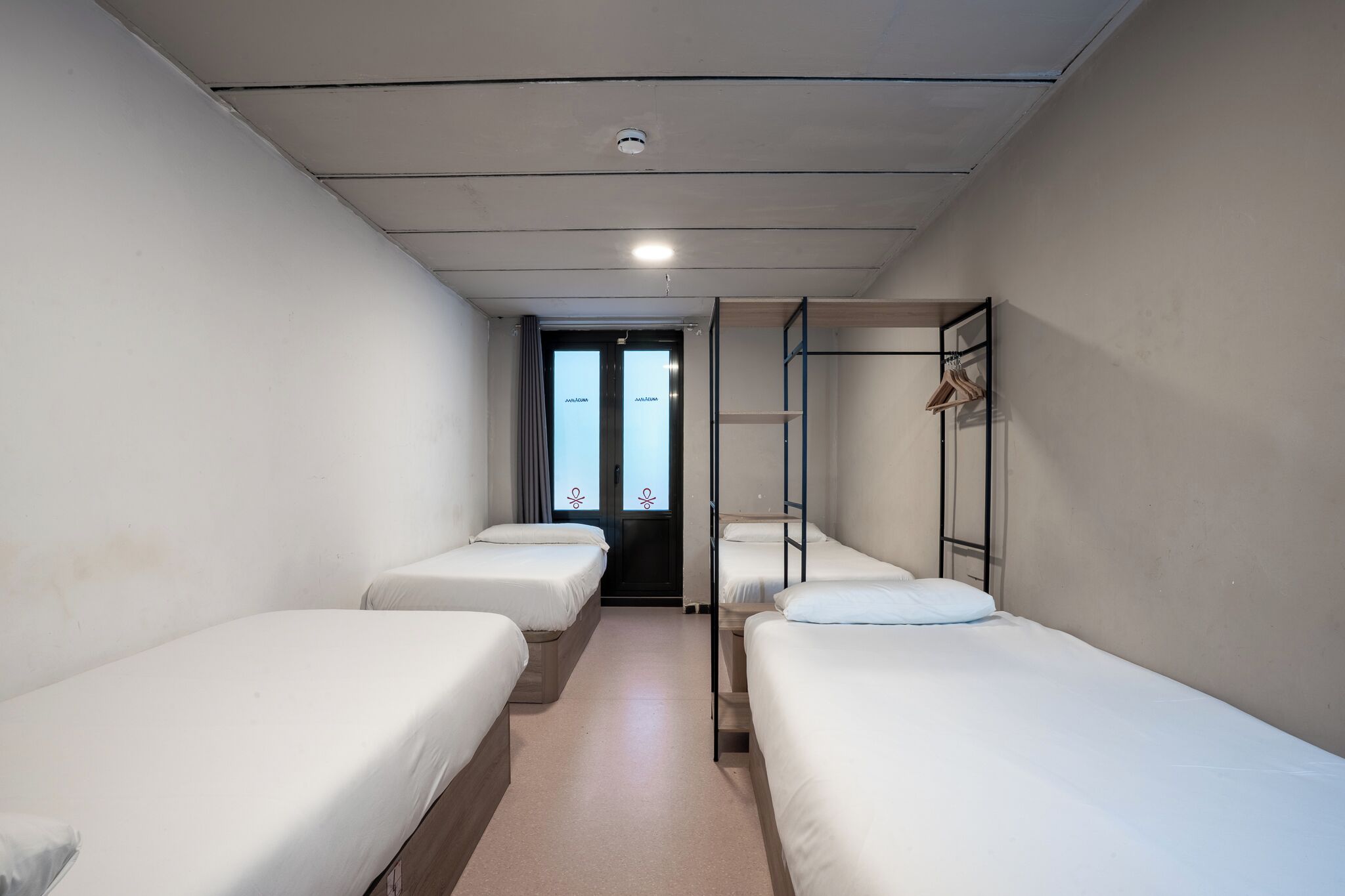 A narrow corridor with four single beds aligned along the walls, a door at the end, and a metal ladder against the wall.