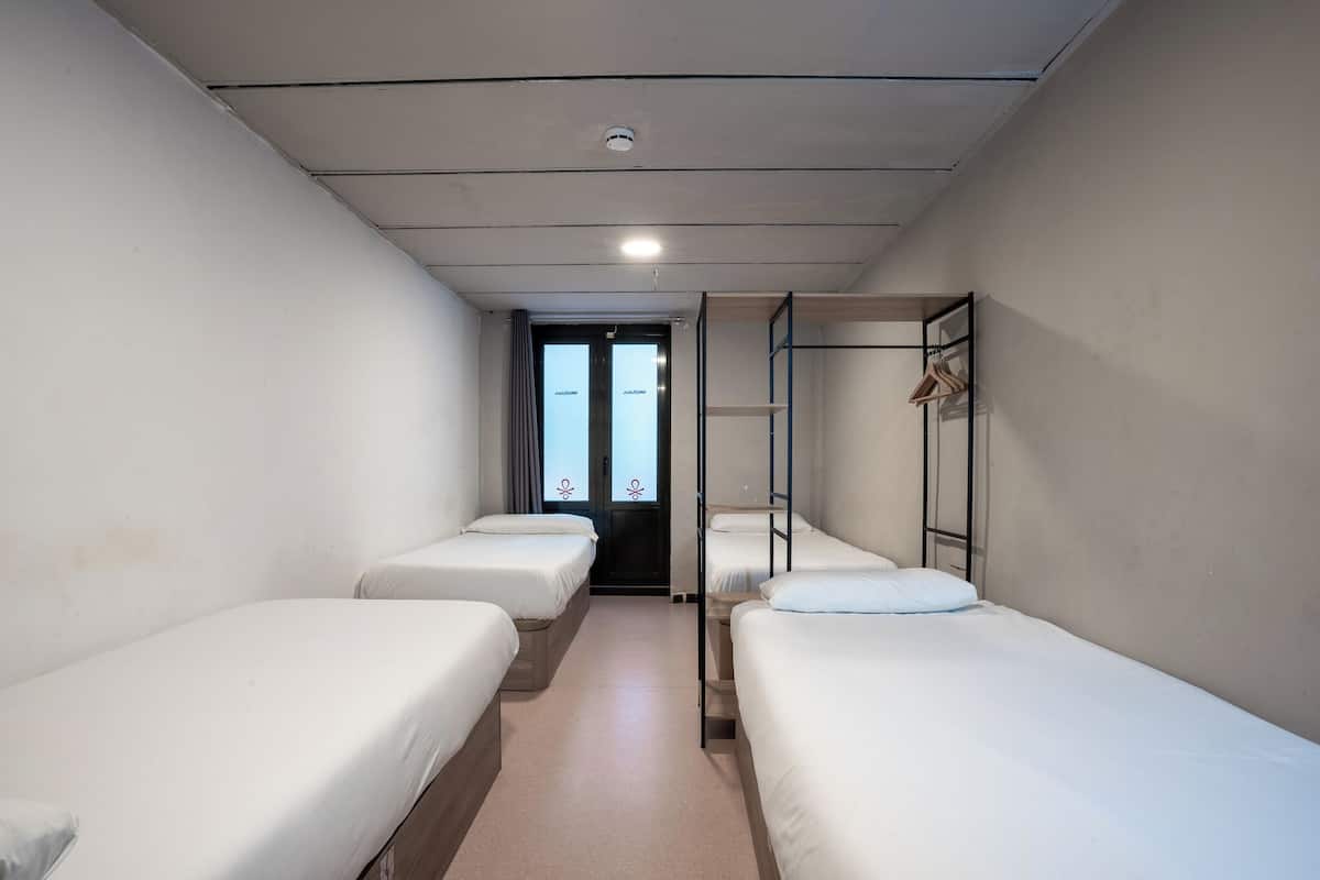 A narrow corridor with four single beds aligned along the walls, a door at the end, and a metal ladder against the wall.