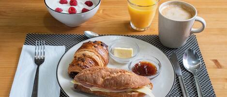 Une table dressée pour le petit-déjeuner avec des croissants, un bol de yaourt aux framboises, un verre de jus d’orange, une tasse de café et une assiette avec du beurre et de la confiture.