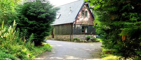 Un chalet rustique avec un toit en pente, entouré d’une végétation luxuriante et un chemin de gravier menant à l’entrée.