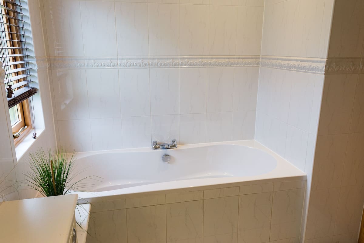 Un baño con bañera, paredes de azulejos blancos, una ventana con persianas y una planta en maceta.