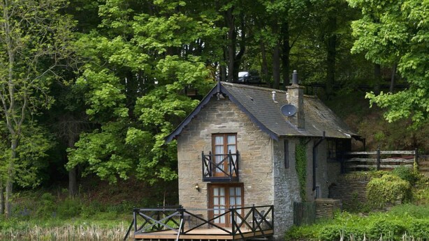 A stone cottage with a wooden deck, surrounded by lush greenery and a calm lake. - Forbes of Kingennie Country Resort (Dundee)