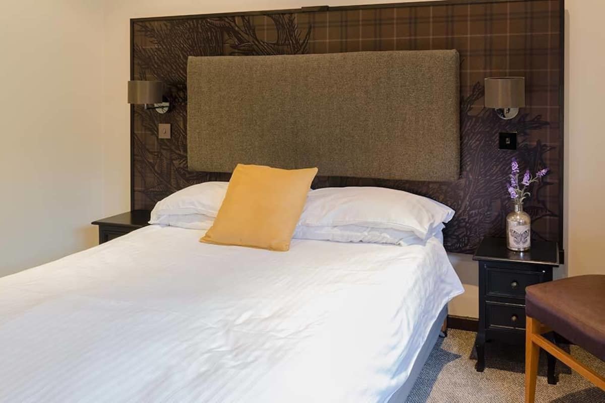 A neatly made bed with white linens and a single tan pillow, a dark headboard, and a small bedside table with a lamp and a vase with flowers.