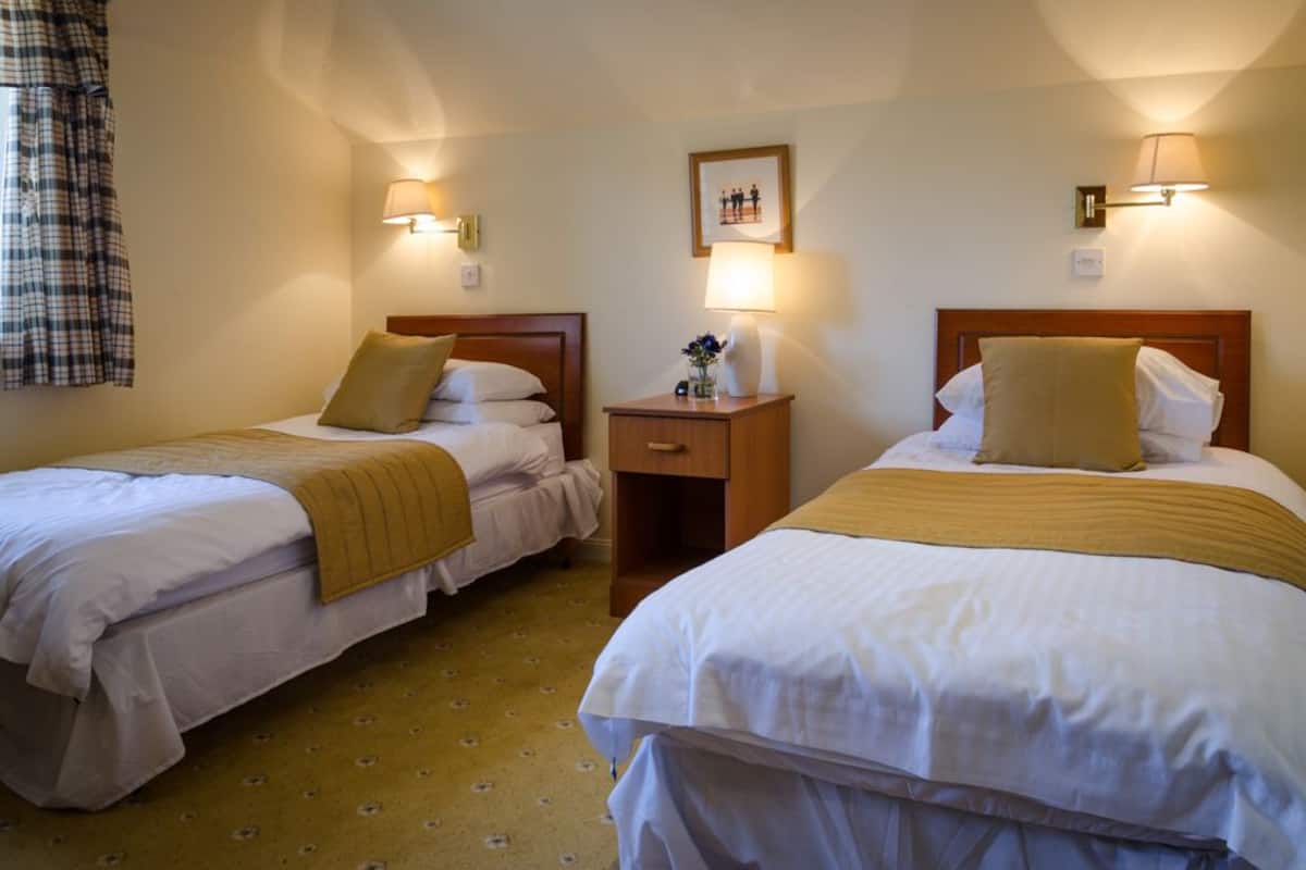 A hotel room with two single beds, each with a wooden headboard, white bedding, and a mustard-colored throw blanket. There are bedside lamps, a small table with a vase of flowers, and a framed picture on the wall.