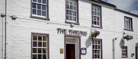 A white building with the sign "THE THRUMS" and a welcoming entrance.