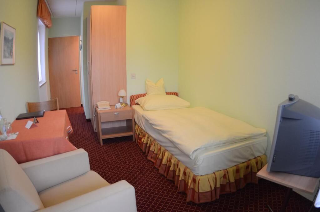 A small hotel room with a bed, a desk with a chair, a television, and a small table with a chair.