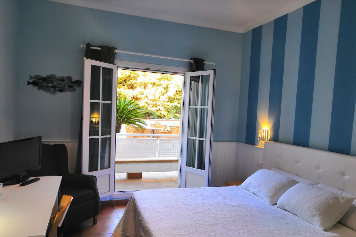 A bedroom with a bed, a desk, a chair, a TV, and a balcony with a view of trees.