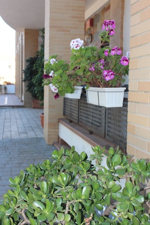 Een gebouw met een bakstenen gevel, een overdekt terras met een bankje en bloeiende planten in potten.