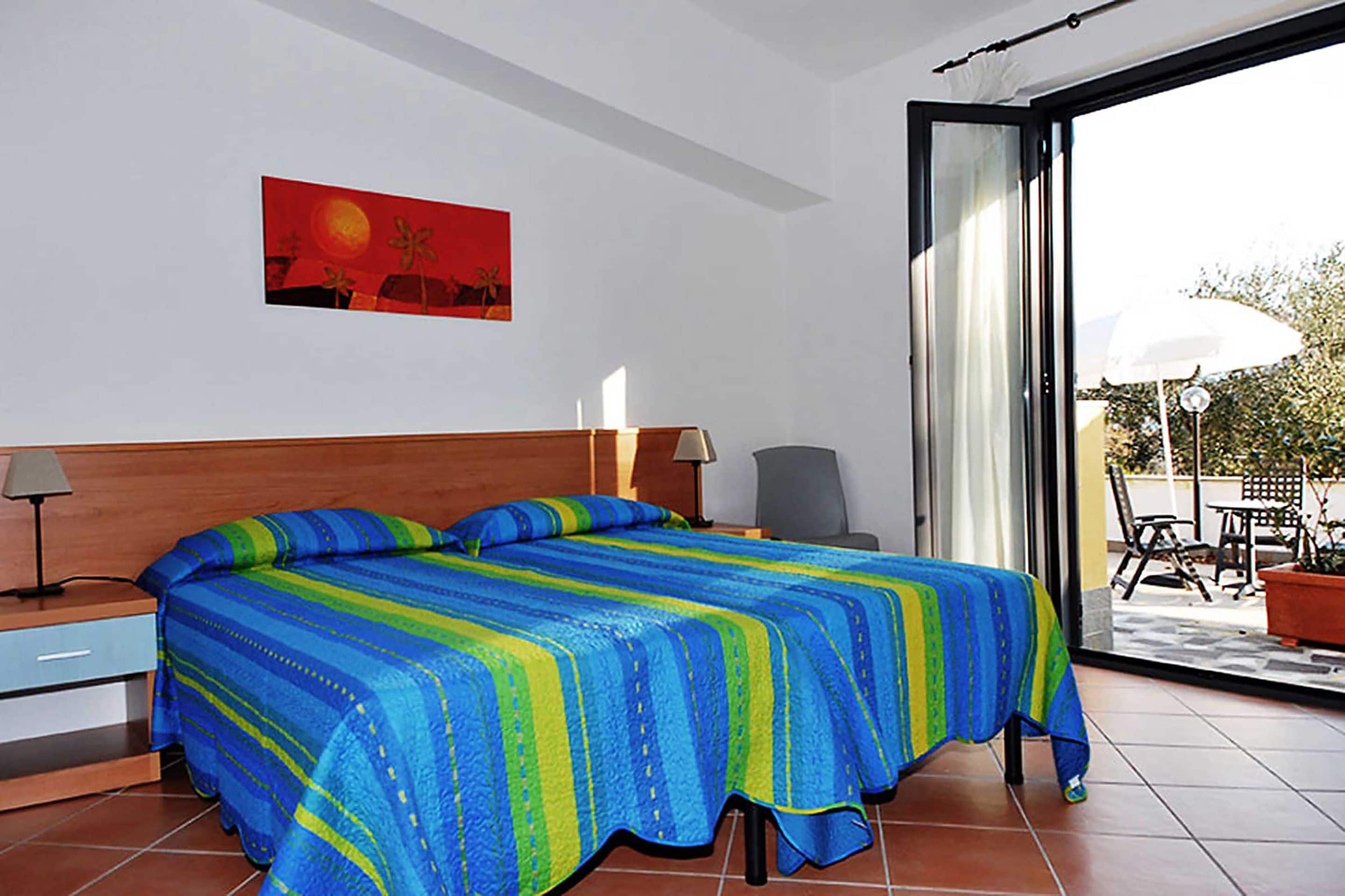 A bedroom with a bed, a nightstand, a lamp, and a window with a view of the outside.