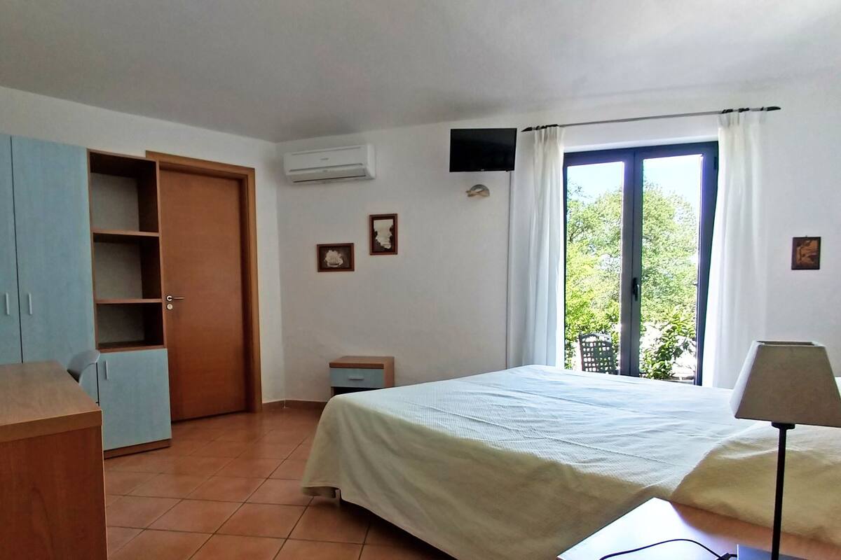 A bedroom with a bed, a desk, a wardrobe, a window with curtains, and a wall-mounted air conditioner.
