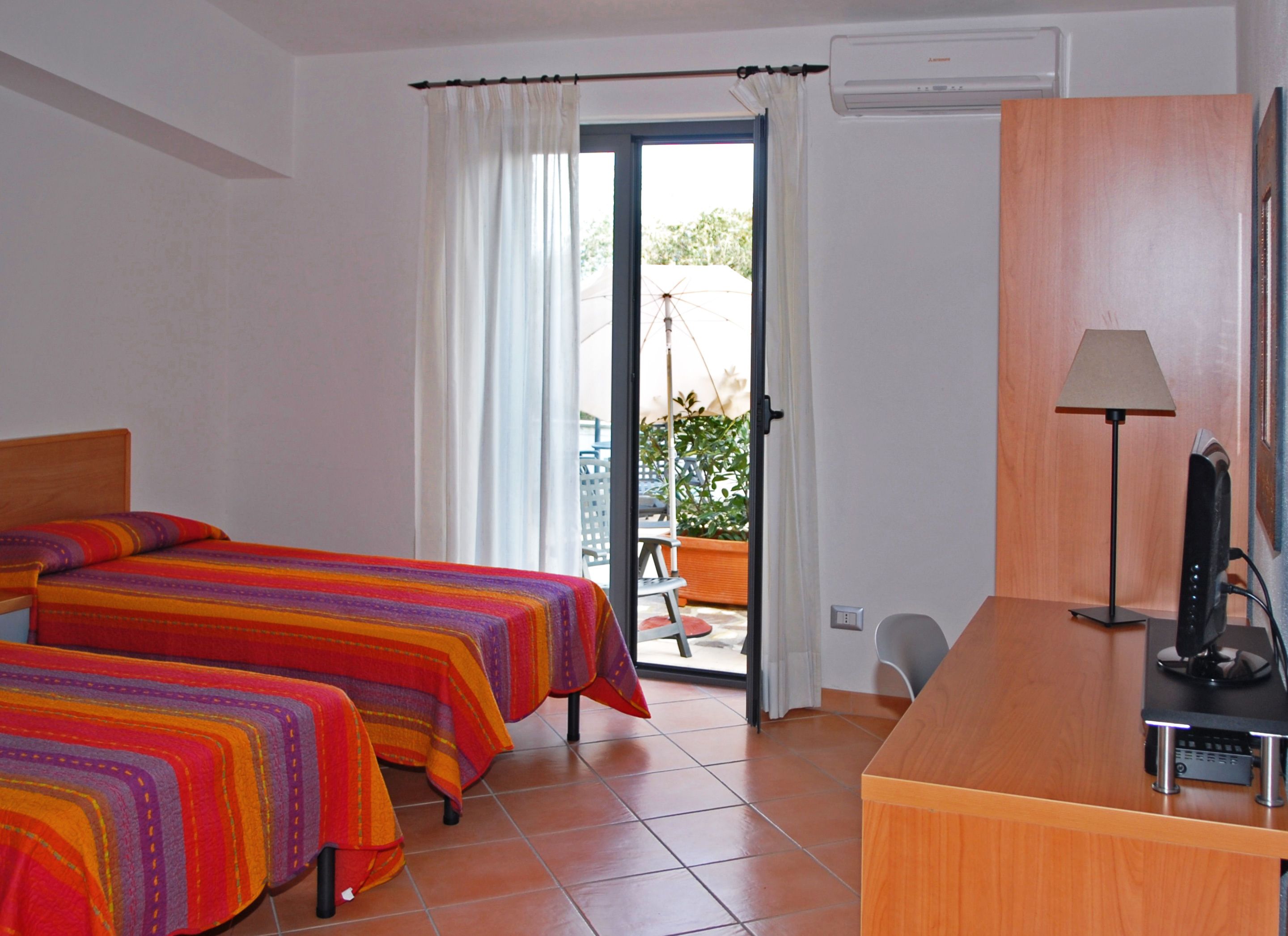 A hotel room with two beds, a desk, a lamp, and a view of the outside.