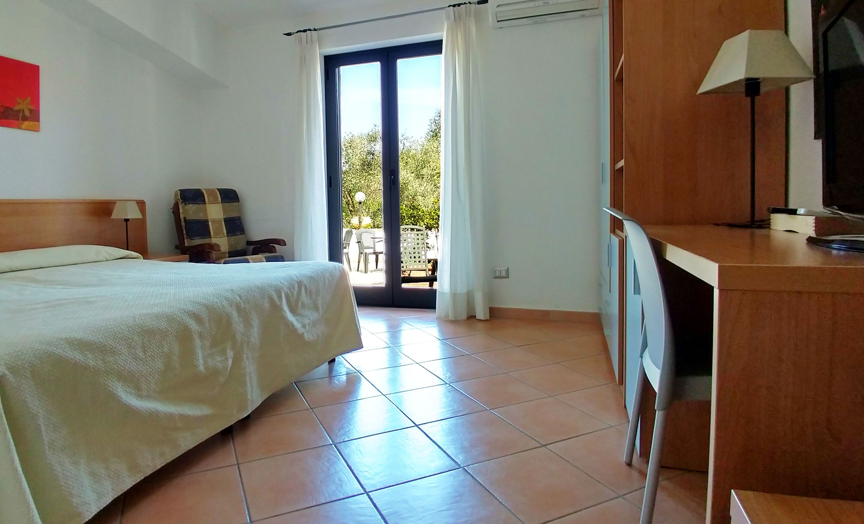 A hotel room with a bed, a desk, a chair, and a view of the outdoors through a glass door.