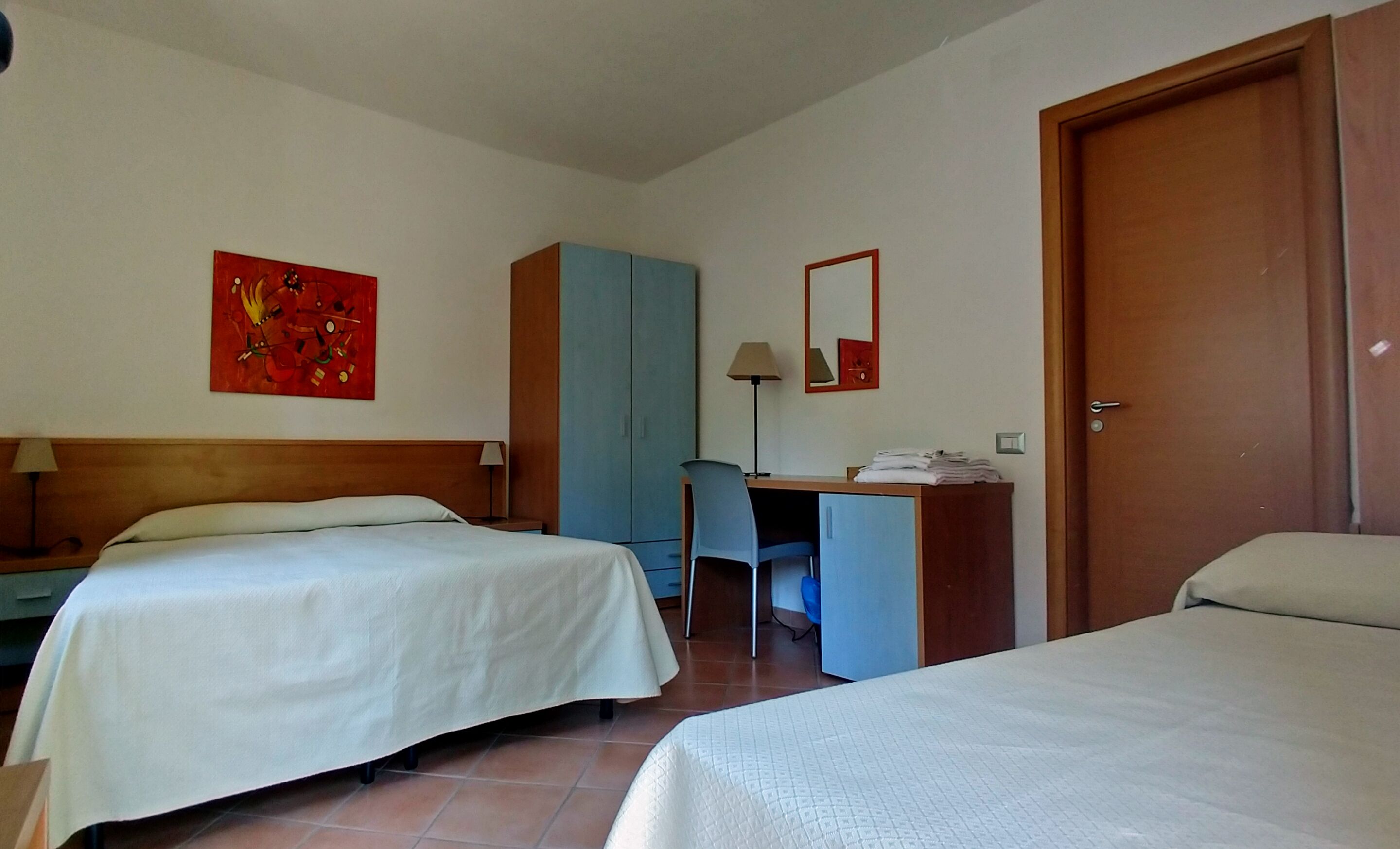 A hotel room with two beds, a desk, a chair, a wardrobe, a lamp, and a painting on the wall.