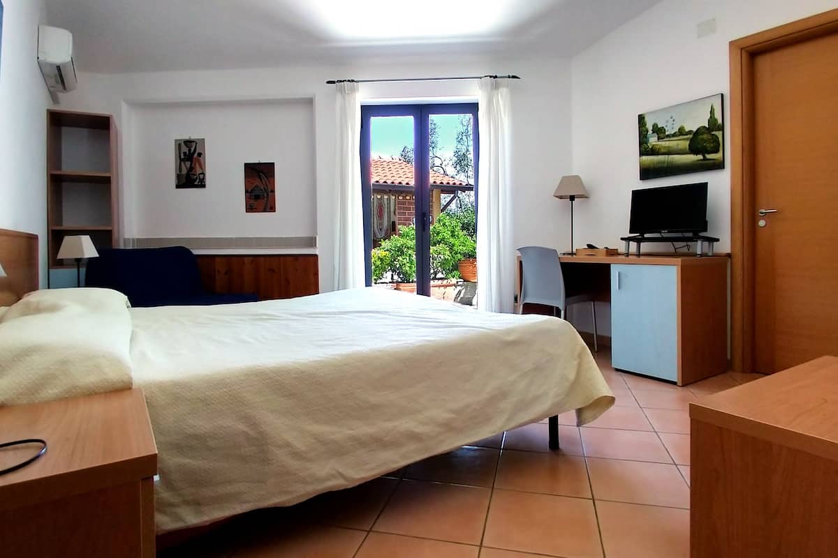 A hotel room with a bed, a desk with a computer, a TV, a chair, a wardrobe, and a window with curtains.