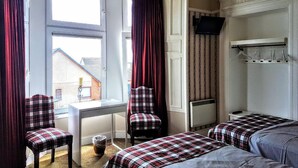 A room with two beds, a desk, and chairs. There is a window with curtains, a radiator, and a TV mounted on the wall. - Seaview Guest House (Carnoustie)