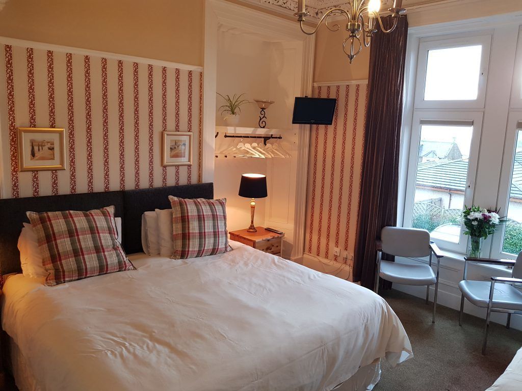 Ein Hotelzimmer mit einem großen Bett, einem Nachttisch mit Lampe, einem an der Wand befestigten Fernseher, einem Sessel und einem Fenster mit Vorhängen.