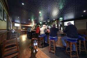 Un bar avec des tabourets en bois, un chien allongé sur le sol et des clients assis au comptoir.