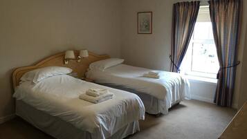 Habitación de hotel con dos camas individuales, cabecera de madera, mesita de noche pequeña, ventana con cortinas y un cuadro enmarcado en la pared.