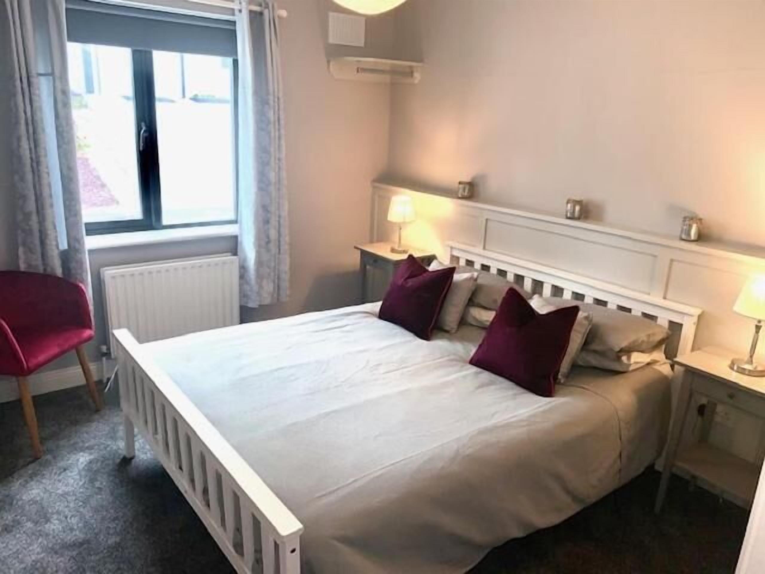 A bedroom with a white bed, two nightstands with lamps, a red chair, and a window with curtains.