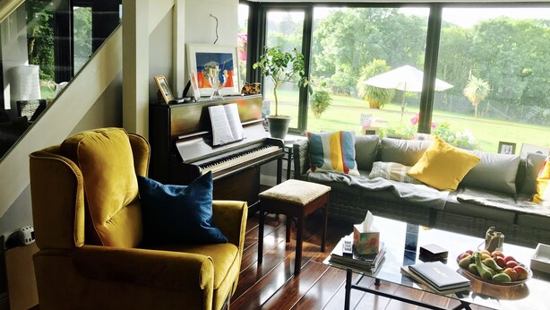 A living room with a piano, a yellow armchair, a sofa with yellow cushions, a coffee table with a fruit bowl, and a view of a garden through large windows. - Railway Country House B&B (Gorey)