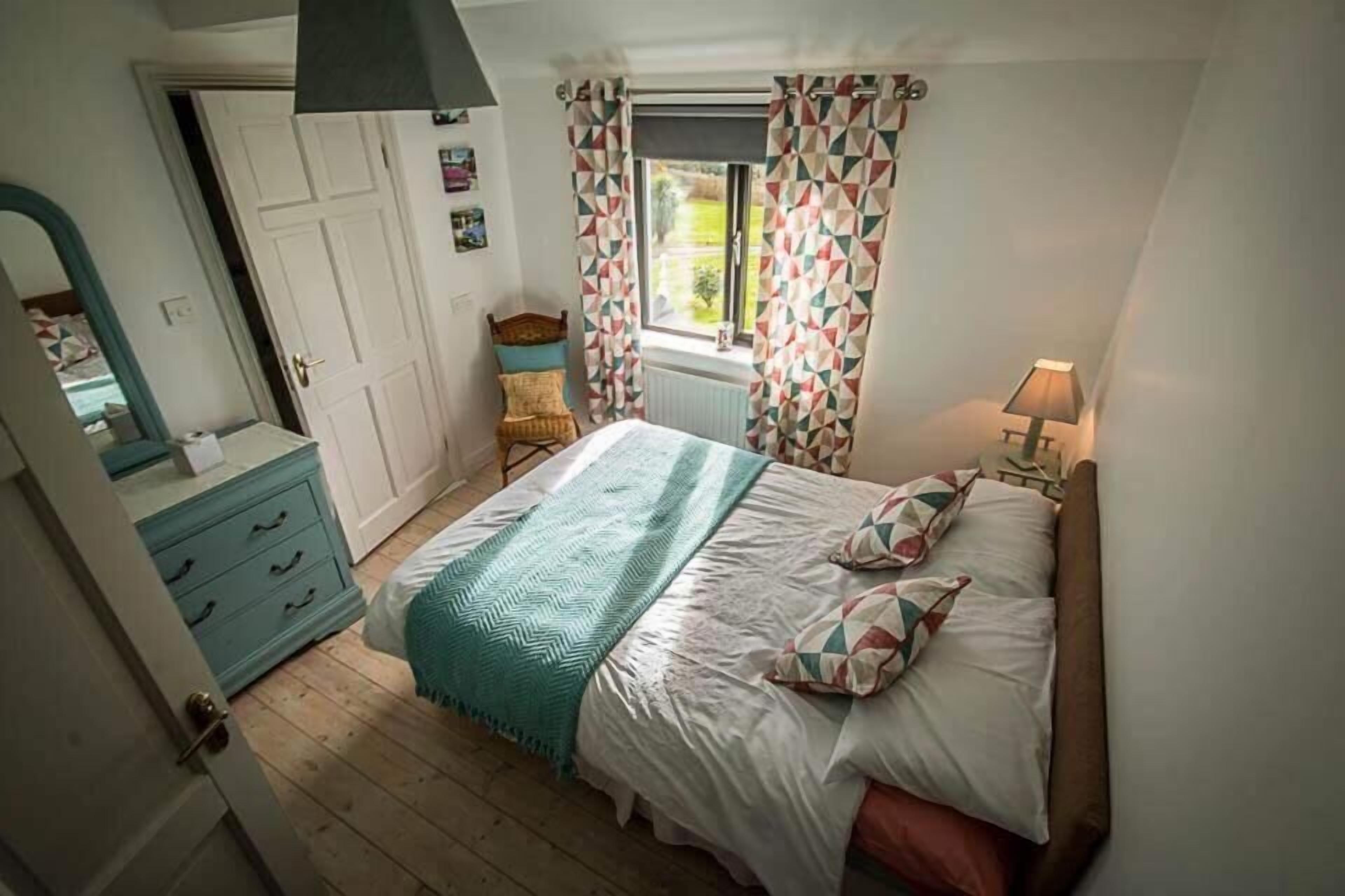 A bedroom with a bed, a dresser, a chair, a window with curtains, and a lamp.