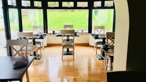 A dining area with wooden flooring, white chairs, and tables. Large windows offer a view of a green lawn. - Railway Country House B&B (Gorey)