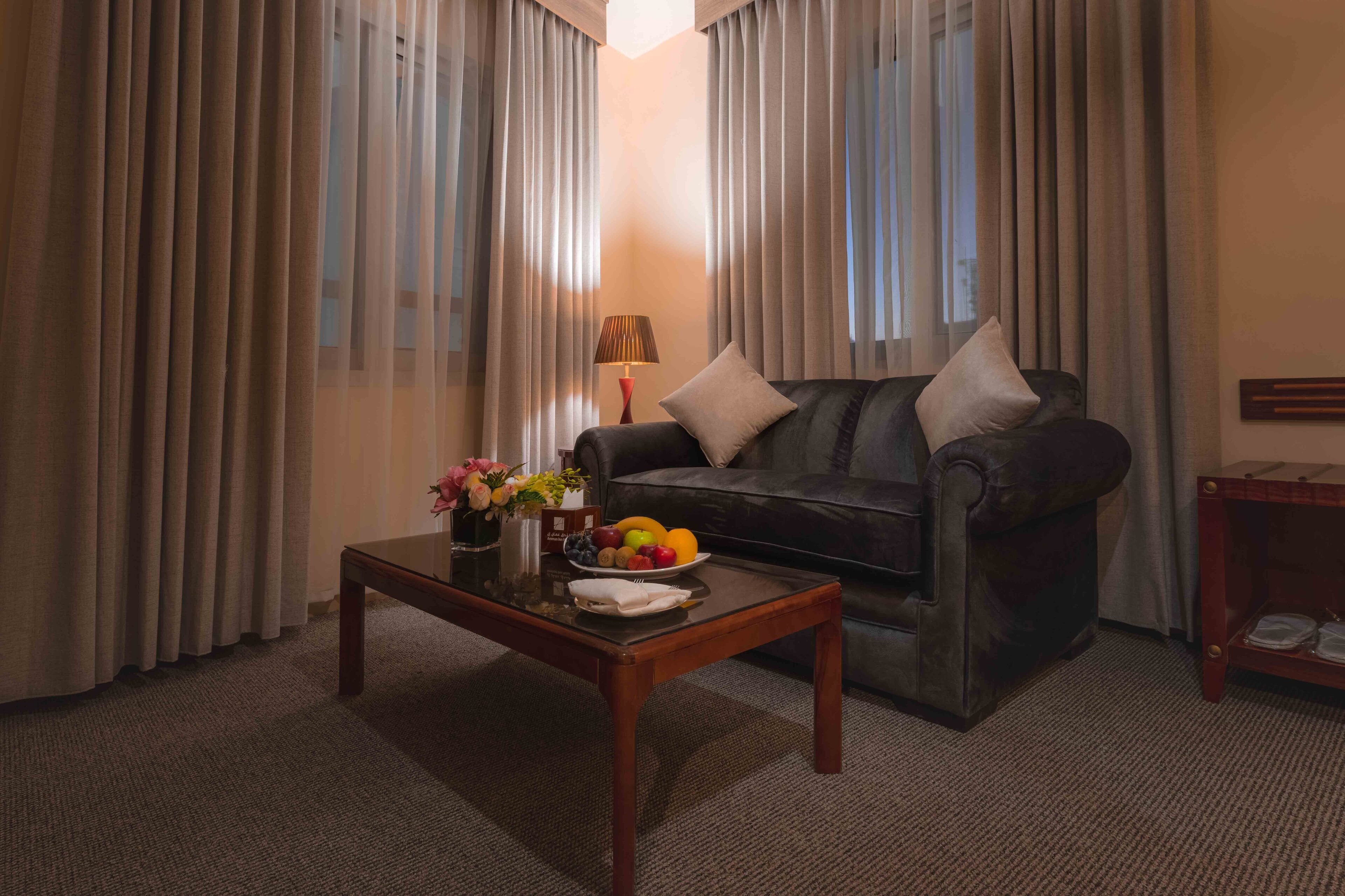 A living room with a dark leather sofa, a glass coffee table with fruit and flowers, and sheer curtains.