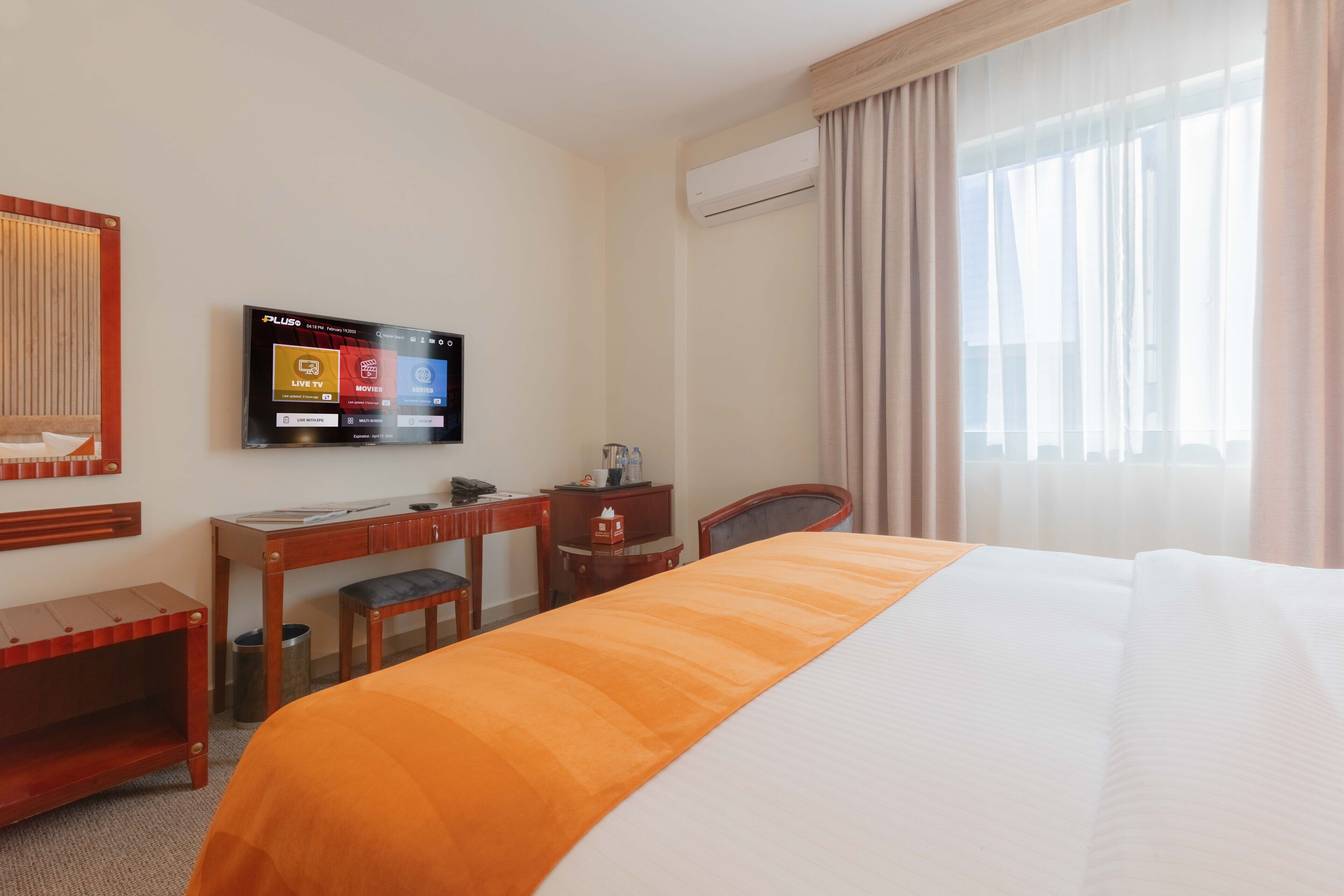 Een hotelkamer met een bed, een bureau, een stoel, een televisie waarop een menu staat en een airconditioning.