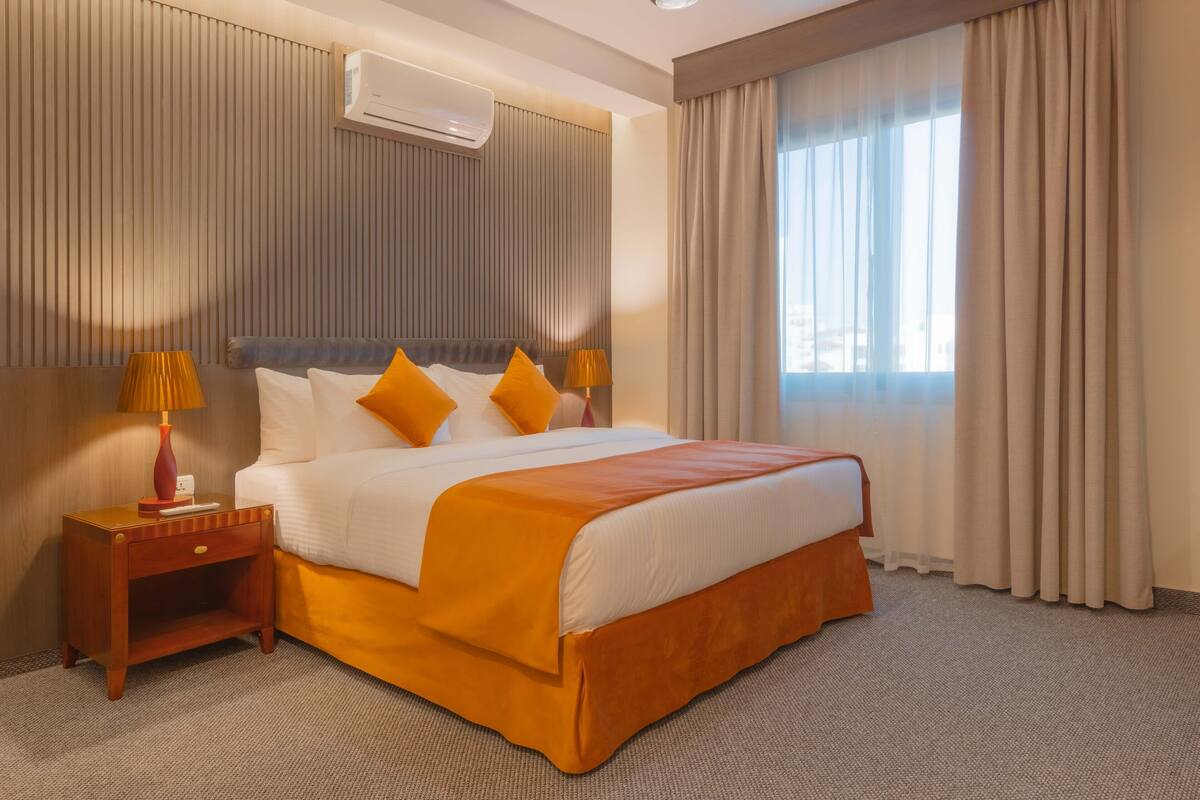 A hotel room with a large bed, a nightstand with a lamp, a wall-mounted air conditioner, and a window with curtains.
