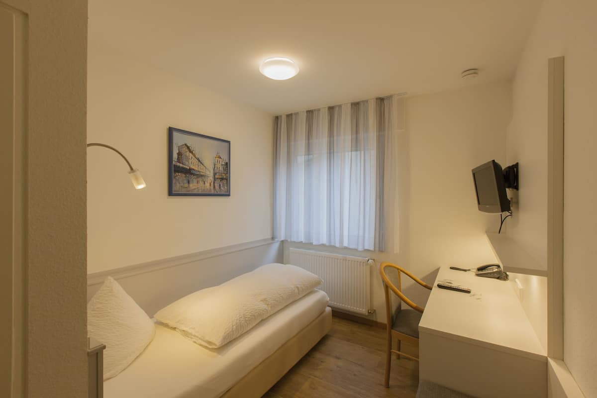 Een kleine, verzorgde slaapkamer met een enkel bed, een bureau, een stoel, een wandgemonteerde televisie en een raam met gordijnen.