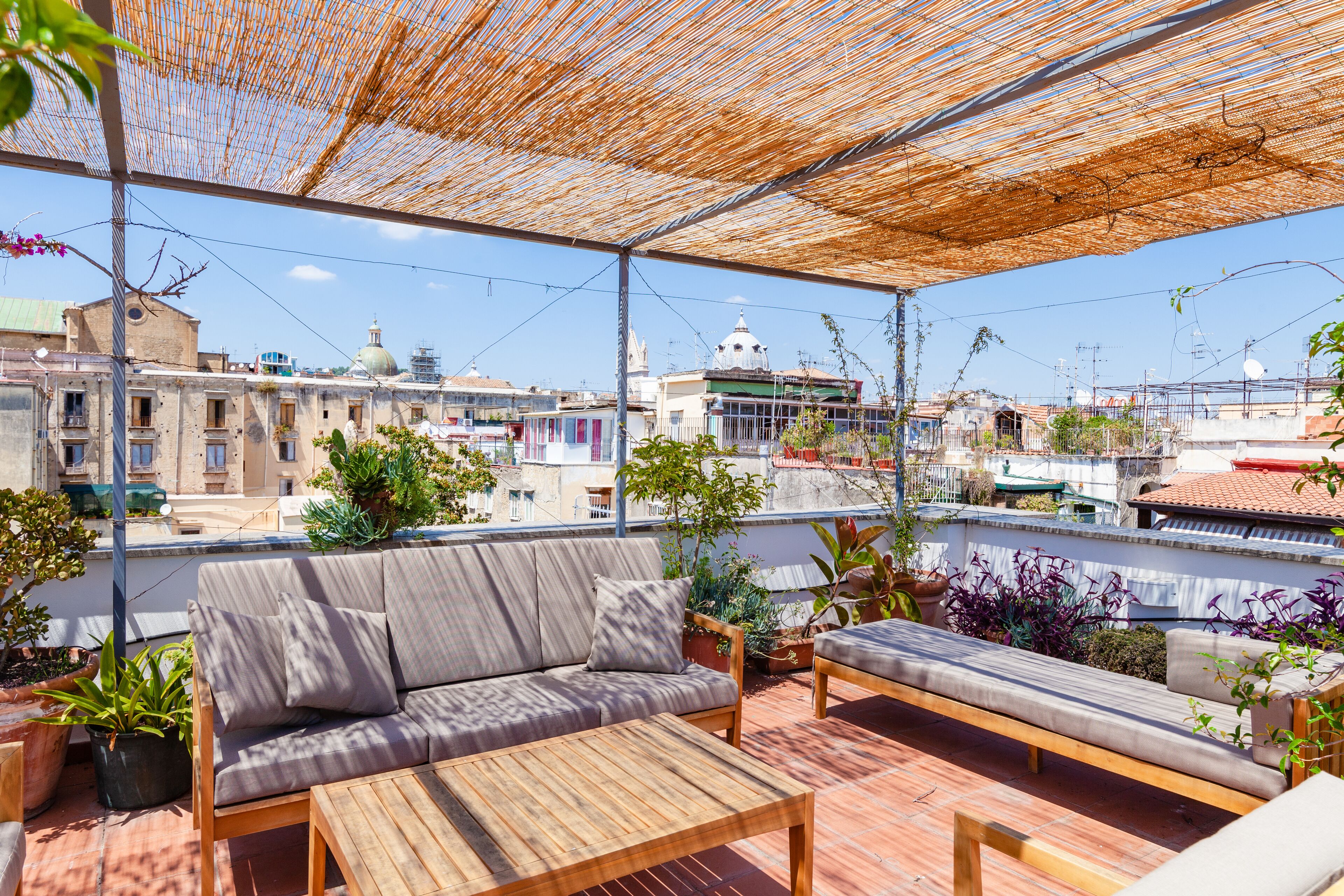 A rooftop terrace with a wooden table and two sofas, surrounded by potted plants and a bamboo canopy.