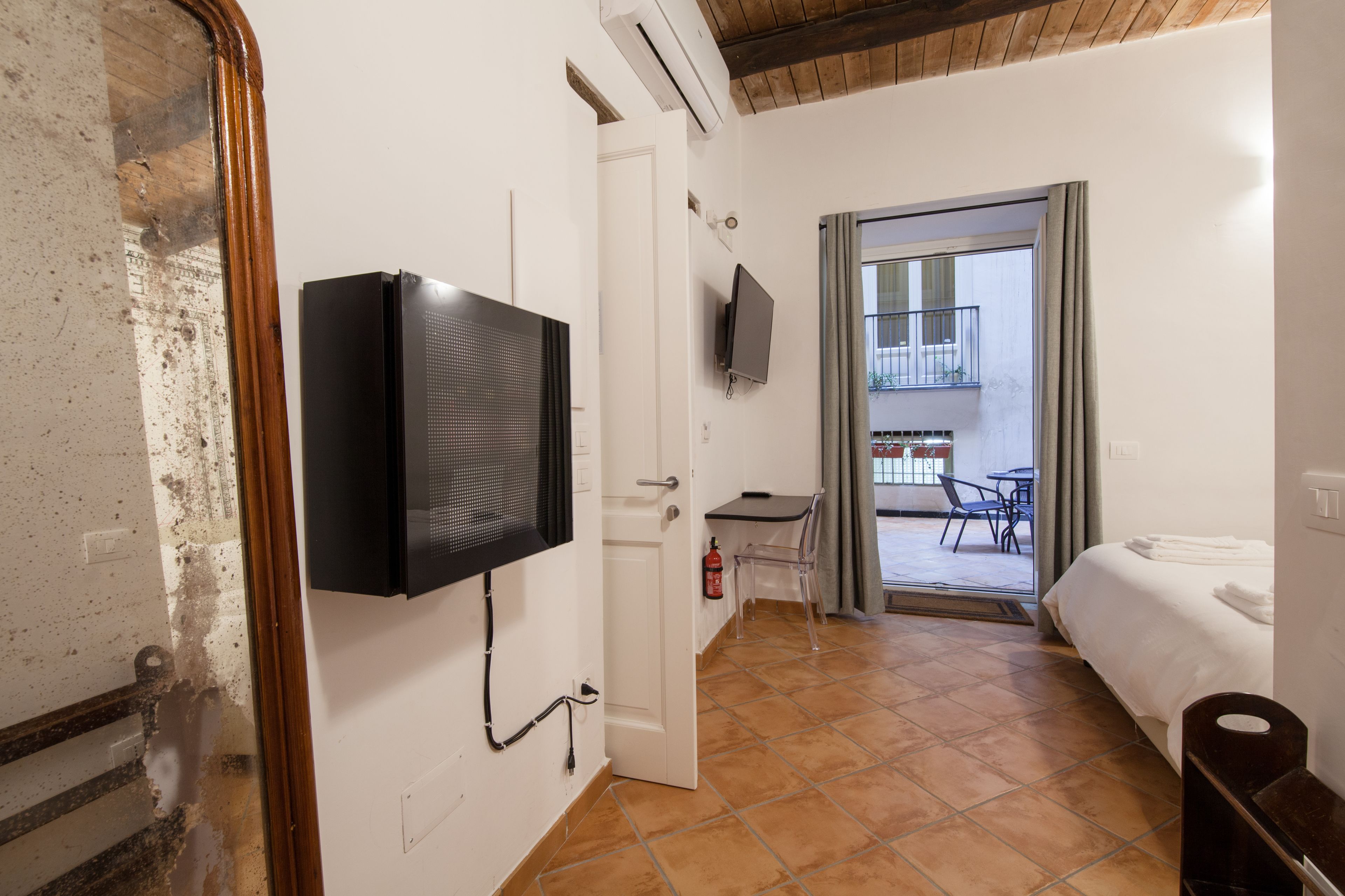 A hotel room with a television mounted on the wall, a bed, a small table with a chair, and a balcony with a view.