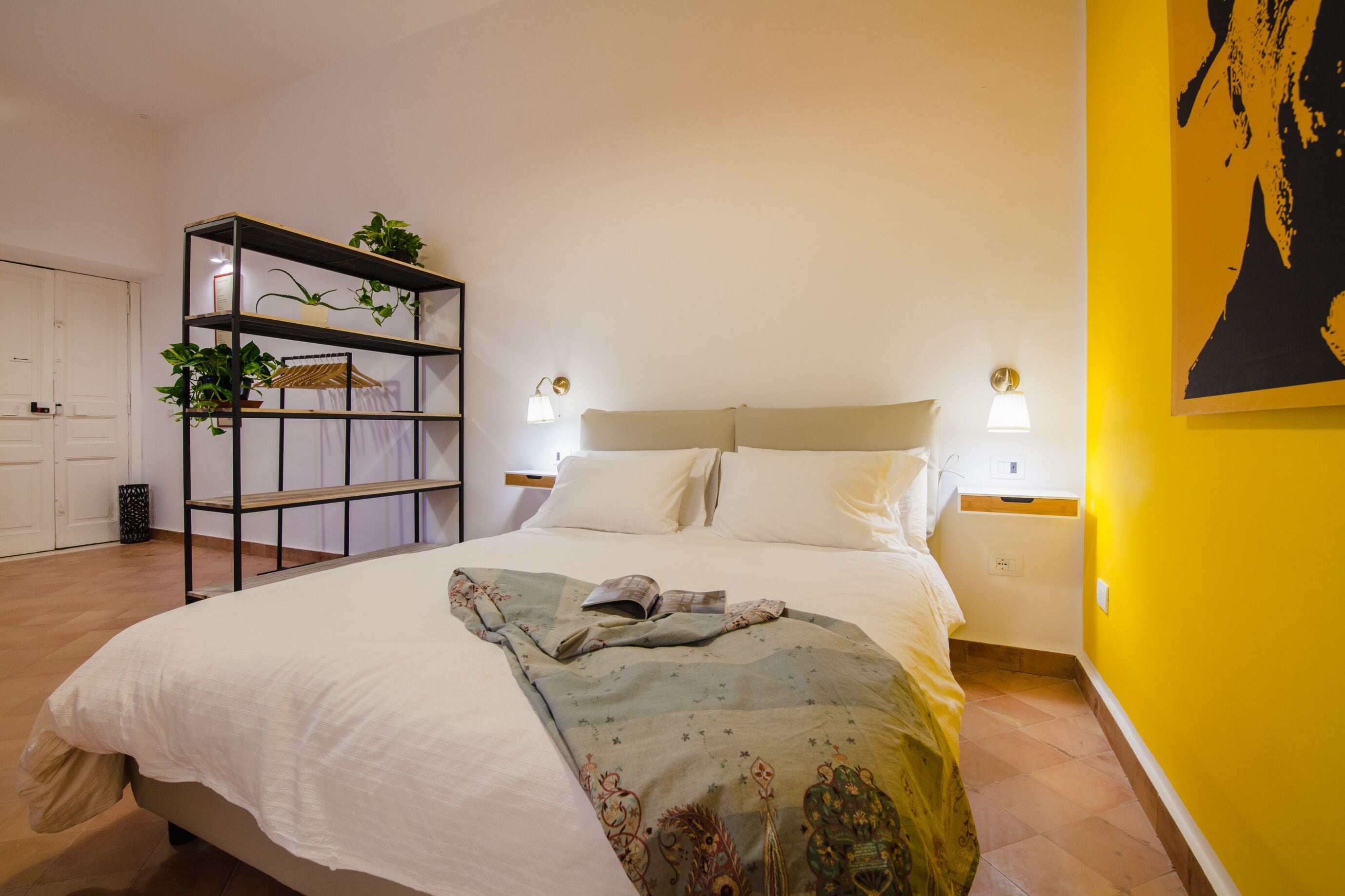 A bedroom with a bed, a bedside table, a shelf with plants, and a wall with a painting.