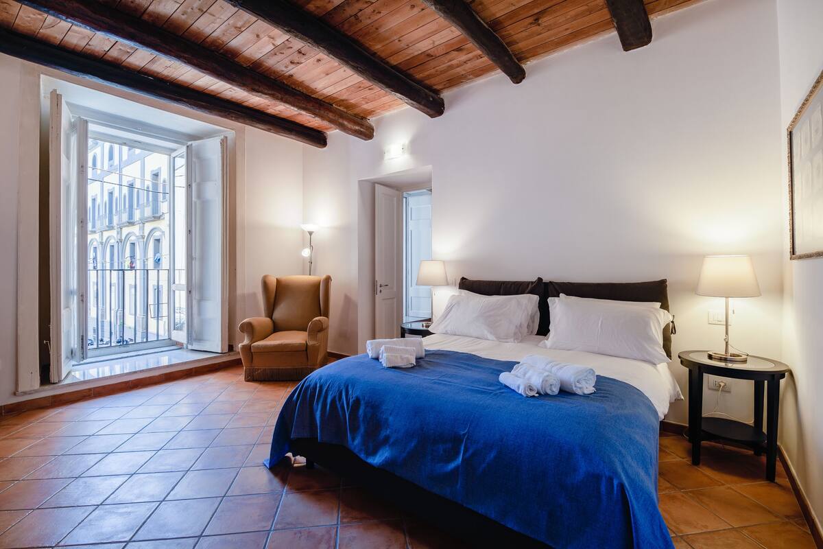 A hotel room with a large bed, a chair, a nightstand, and a view of the outside.