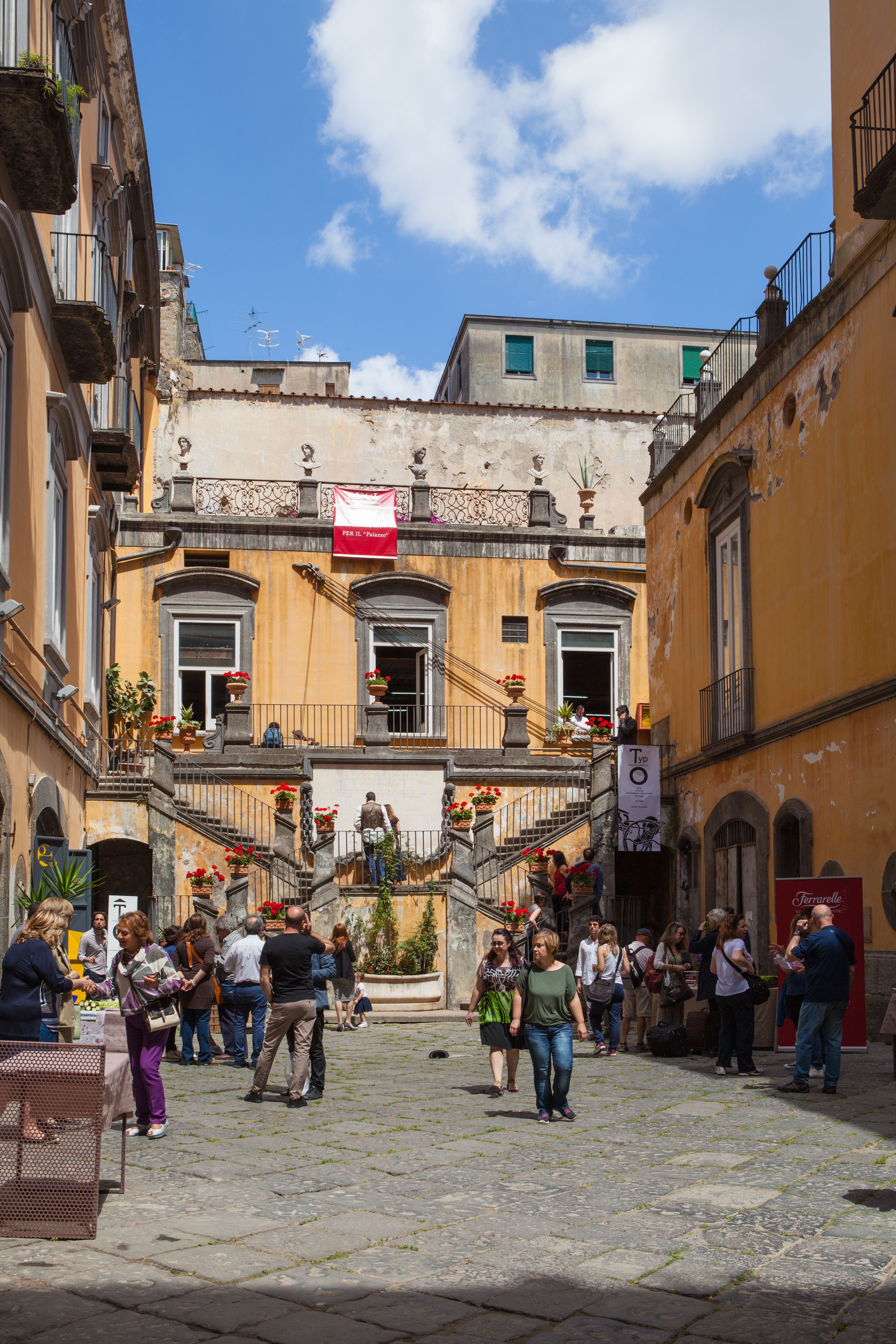 A bustling street scene with a building featuring a central staircase, balconies with potted plants, and a red banner.