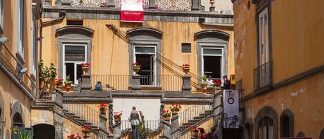 A bustling street scene with a building featuring a central staircase, balconies with potted plants, and a red banner.