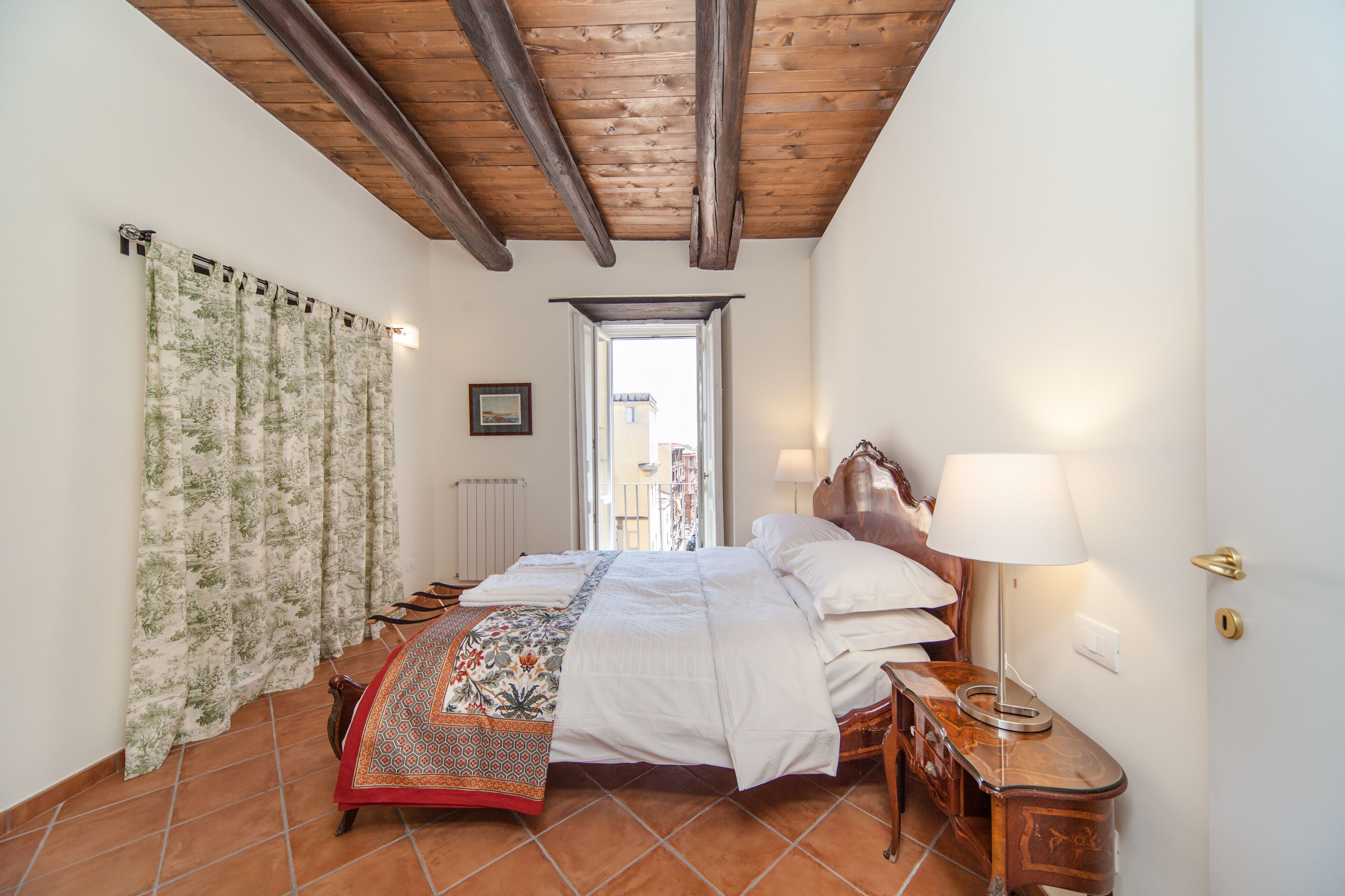 A bedroom with a wooden ceiling, a bed with a patterned bedspread, a nightstand with a lamp, and a view through an open door.