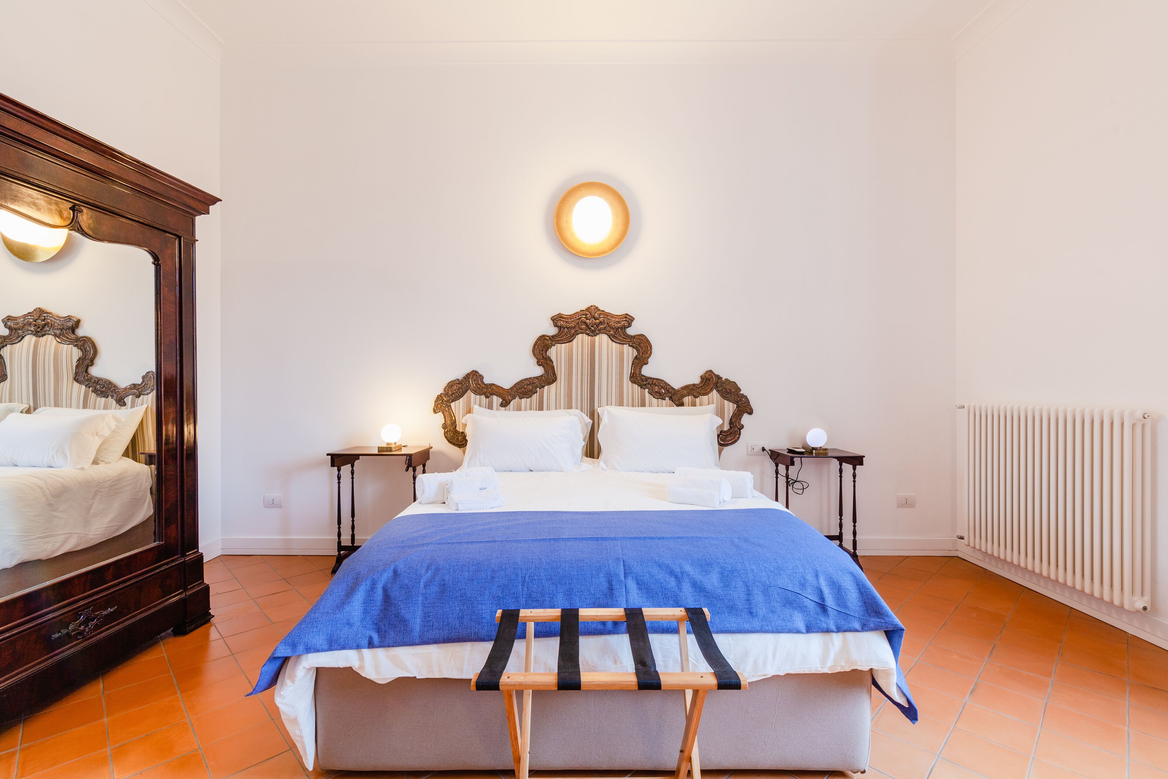Una cama bien hecha con una manta azul, un banco de madera, un espejo grande y una lámpara de pared.