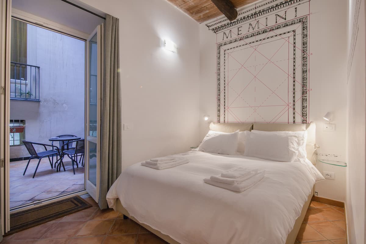 A neatly made bed with white linens, a glass door leading to an outdoor seating area, and a wall with a geometric design.