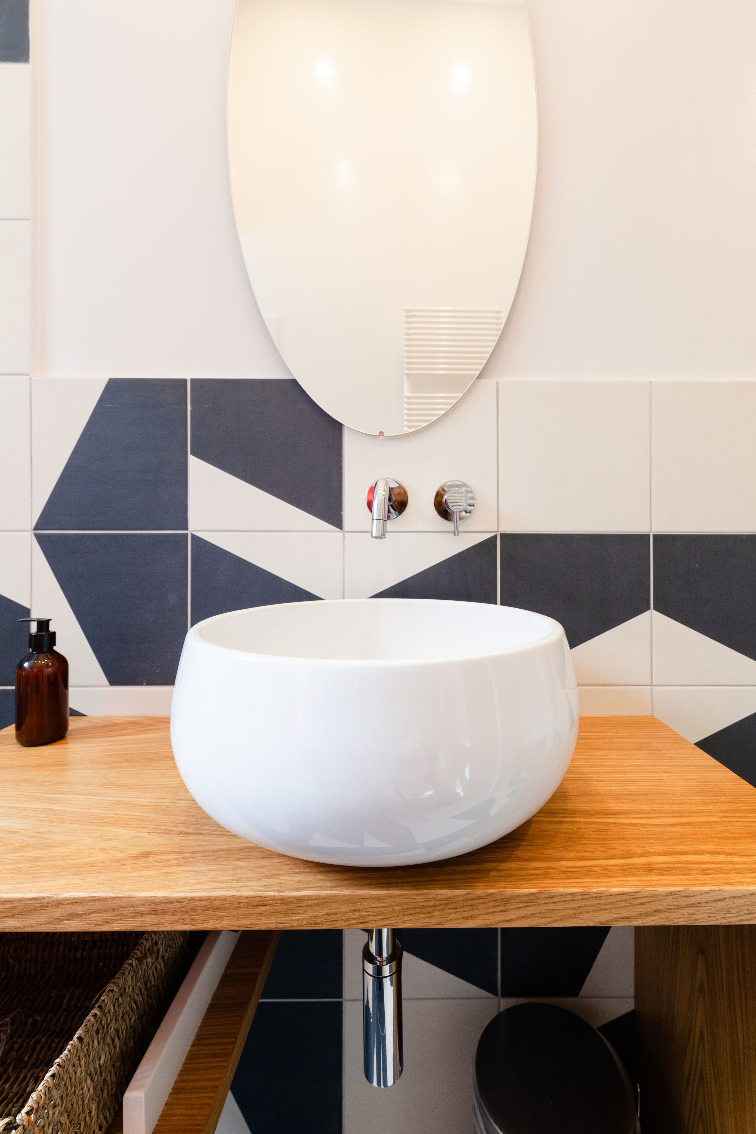 Une salle de bain moderne avec un grand lavabo blanc, un plan de toilette en bois et un revêtement mural en carreaux aux motifs géométriques.