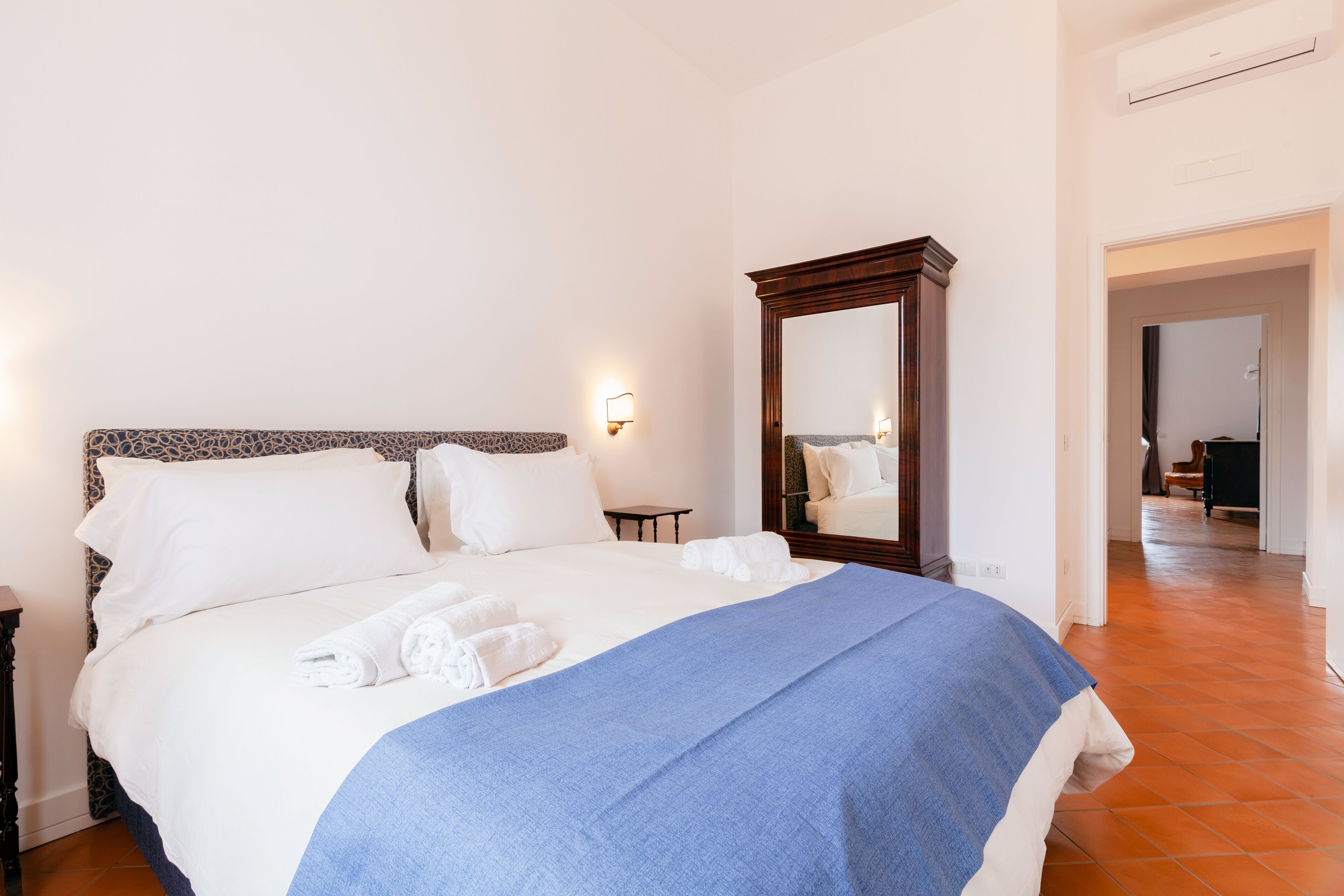 Una cama bien hecha con sábanas blancas y una manta azul, una mesita de noche de madera, una lámpara de pared y un espejo.