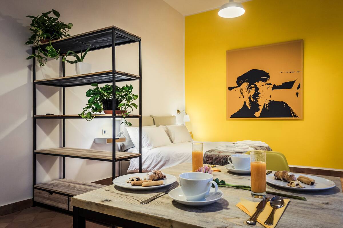 A neatly arranged breakfast table with plates of food, a glass of orange juice, and a cup. A bed with white bedding and a yellow wall with a black and white painting are in the background.