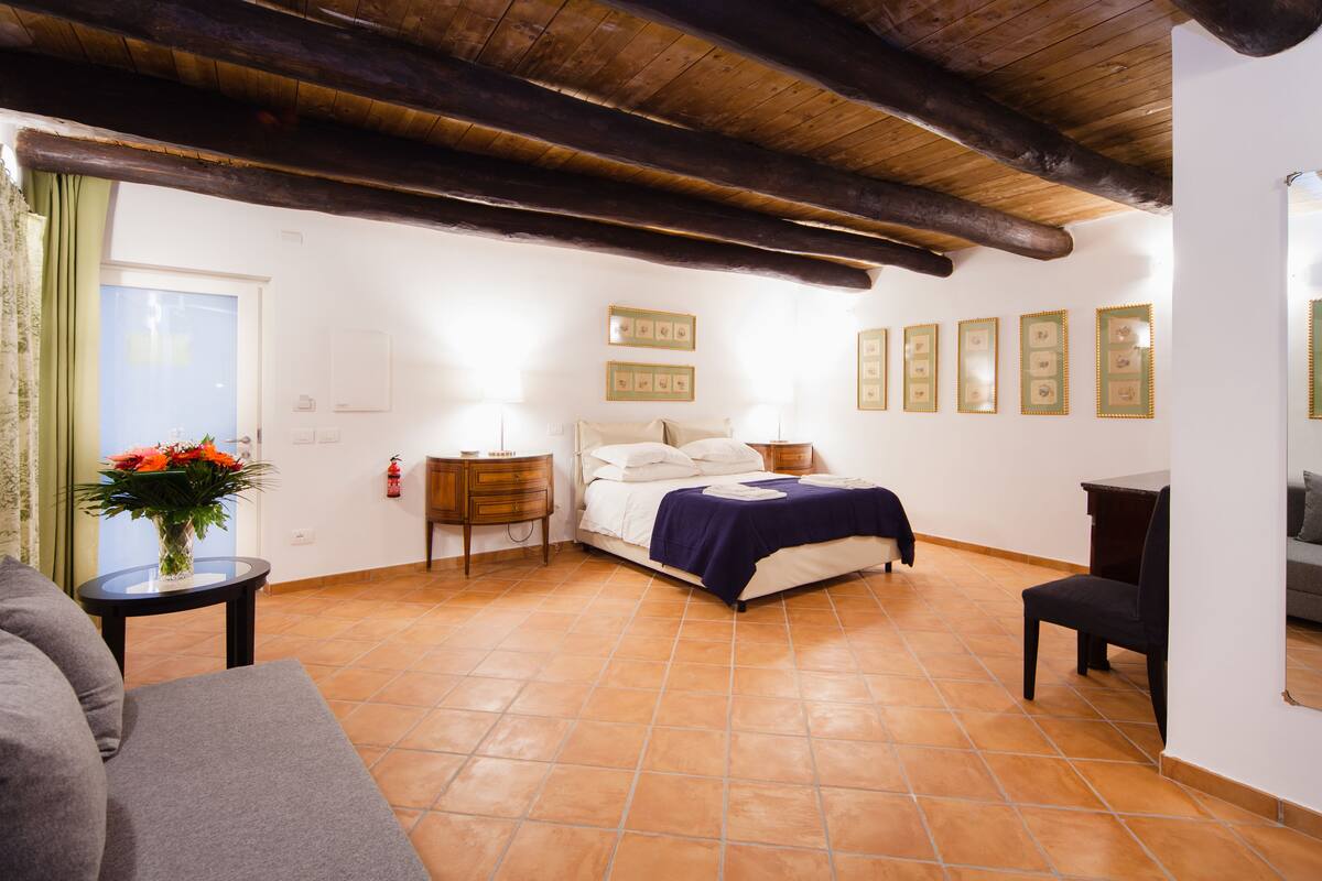 A bedroom with a bed, a nightstand, a chair, a sofa, and a small table with a vase of flowers.