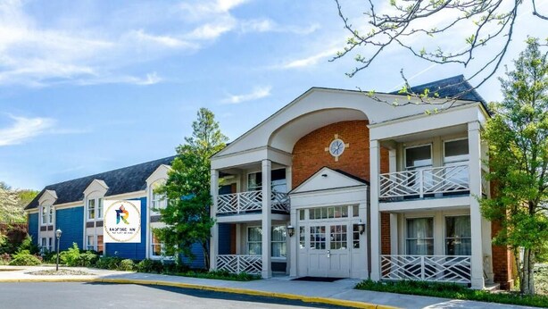Un hotel de varios pisos con un logotipo destacado en la fachada, un balcón en el segundo piso y un estacionamiento al frente.