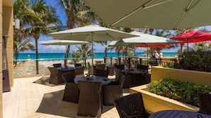 Un patio exterior con mobiliario de mimbre y sombrillas, con vistas a una playa con palmeras y un cielo azul despejado.