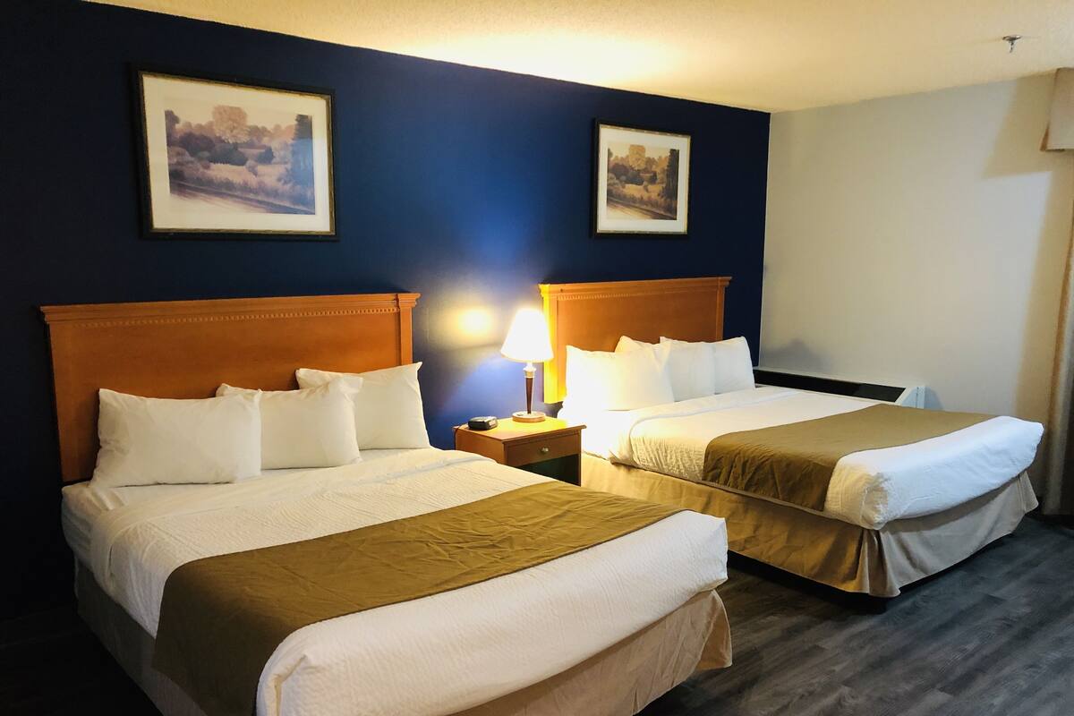 A hotel room with two beds, each with a wooden headboard, white bedding, and a brown throw blanket. There are two framed pictures on the wall, a bedside lamp, and a nightstand with a clock.