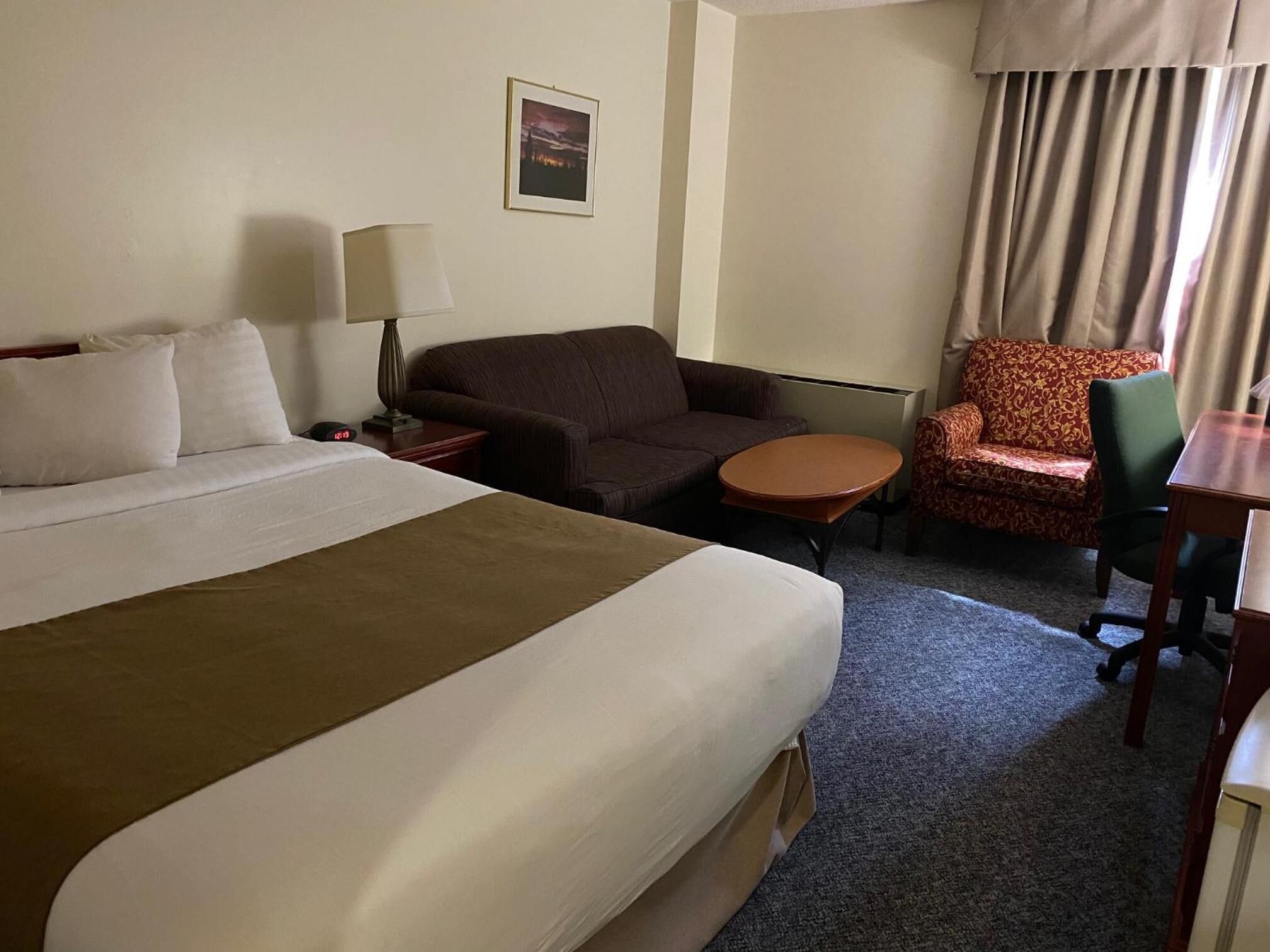 Habitación de hotel con una cama grande, una manta marrón, un sillón marrón, una mesita redonda pequeña, un sillón estampado, una lámpara y un cuadro en la pared.