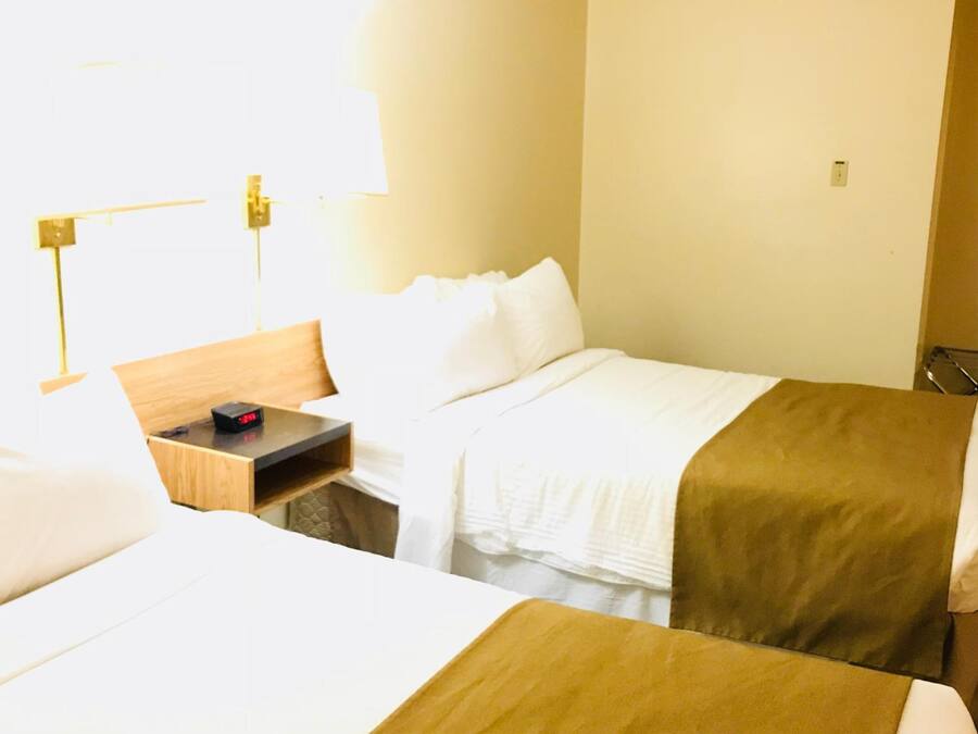 A hotel room with two beds, a bedside table with a clock, a lamp, and a window with curtains.